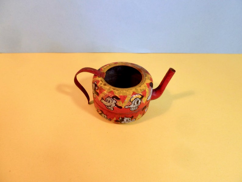 May include: A small, red and yellow metal teapot with a cartoon character design. The teapot has a spout and a handle.