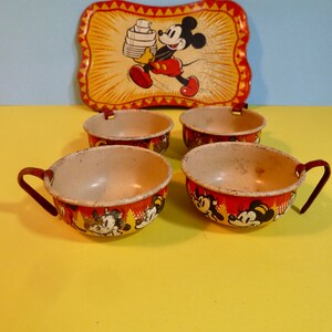 May include: Vintage Mickey Mouse children's tea set with four cups and a tray. The cups are red with a white rim and feature a Mickey Mouse design. The tray is yellow with a red border and features a Mickey Mouse design carrying a stack of plates.