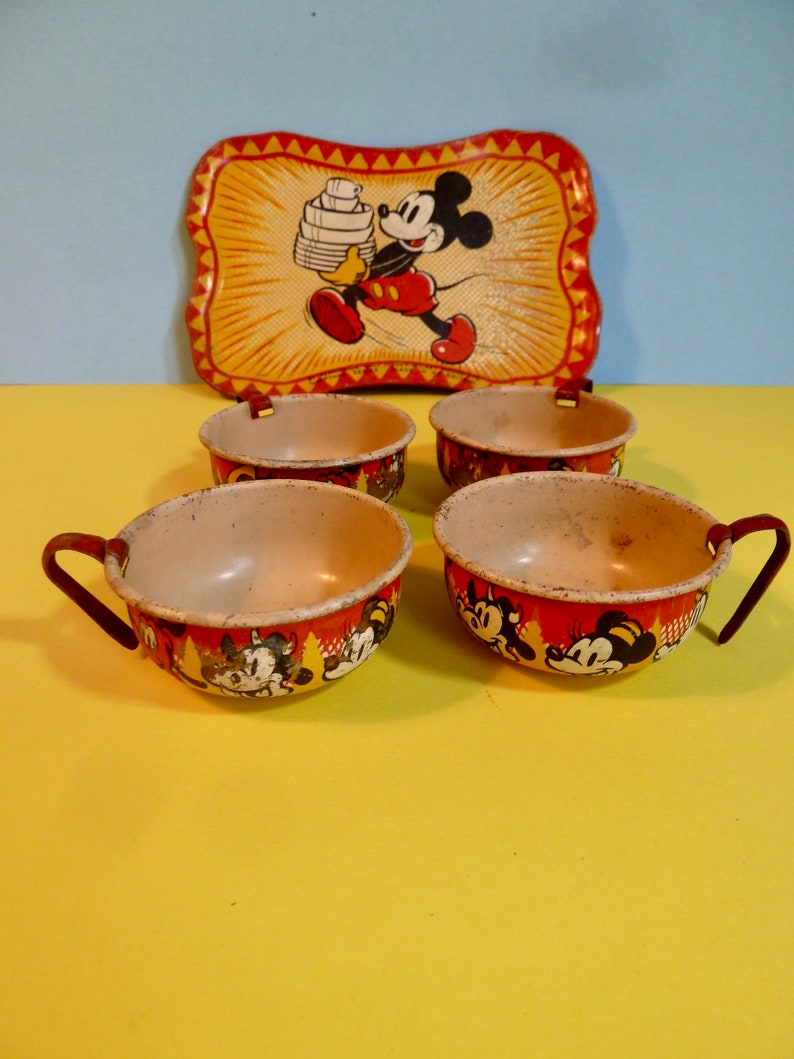 May include: Vintage Mickey Mouse children's tea set with four cups and a tray. The cups are red with a white rim and feature a Mickey Mouse design. The tray is yellow with a red border and features a Mickey Mouse design carrying a stack of plates.
