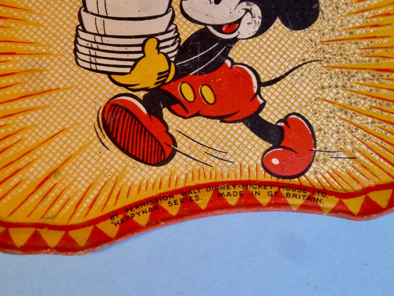 May include: A yellow and red vintage tin plate featuring Mickey Mouse carrying a stack of plates. The text "By Permission Walt Disney-Mickey Mouse Ltd. Happynak Series. Made in Gt Britain." is printed on the bottom of the plate.
