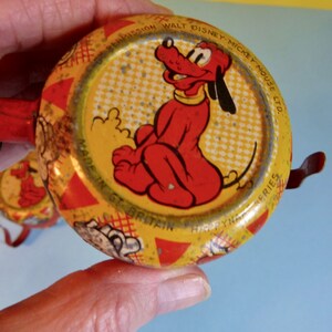 May include: A yellow and red tin toy featuring a picture of Pluto the dog. The toy is made in Great Britain and is part of the "Droopy" series. The text on the toy reads "By Permission Walt Disney-Mickey Mouse Ltd."