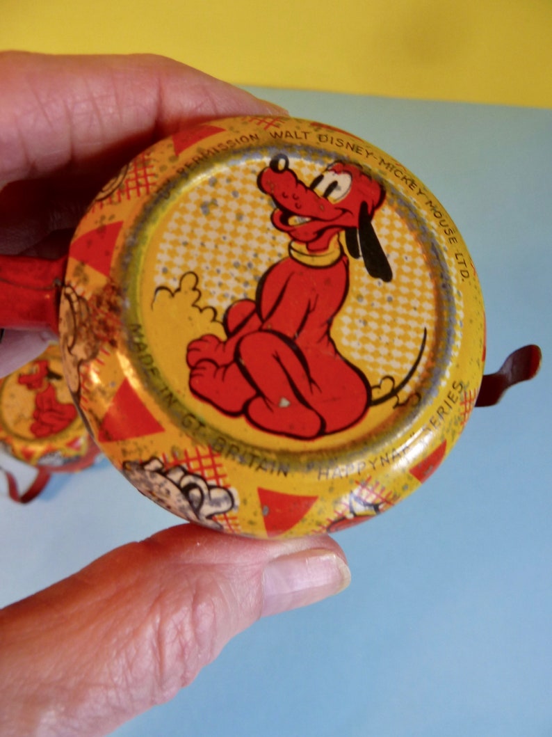 May include: A yellow and red tin toy featuring a picture of Pluto the dog. The toy is made in Great Britain and is part of the "Droopy" series. The text on the toy reads "By Permission Walt Disney-Mickey Mouse Ltd."