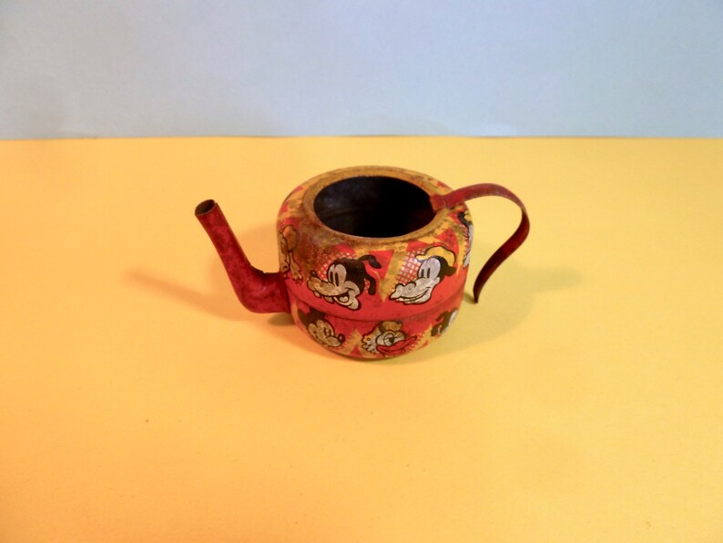 May include: A red and yellow metal watering can with a cartoon character design. The can has a spout and a handle.