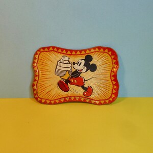 May include: A vintage metal tray featuring a cartoon image of Mickey Mouse carrying a stack of plates. The tray has a yellow background with red and orange accents.