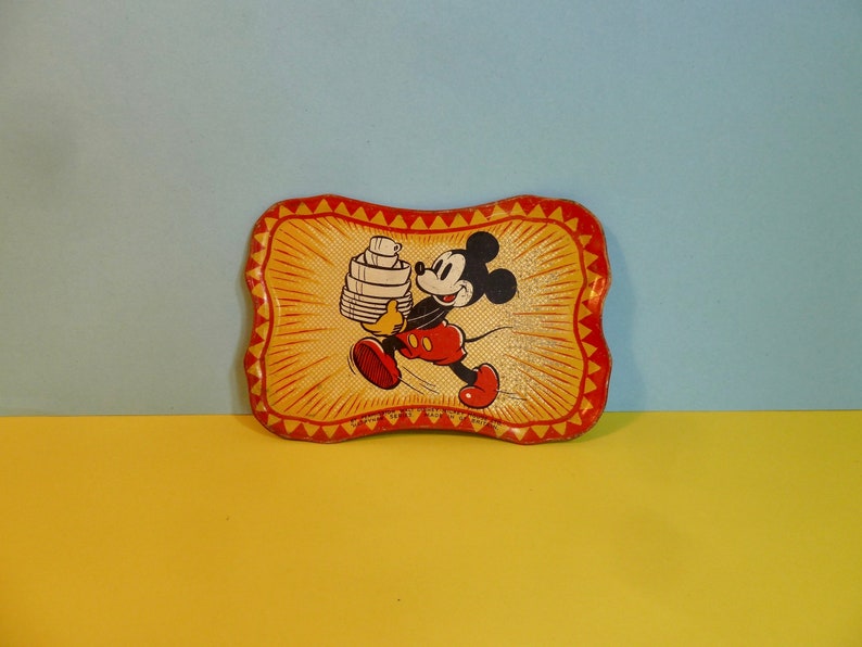 May include: A vintage metal tray featuring a cartoon image of Mickey Mouse carrying a stack of plates. The tray has a yellow background with red and orange accents.