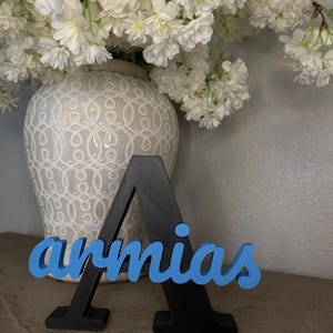 May include: A black wooden letter "A" with the name "armias" in blue script. The letter is standing on a surface in front of a white vase filled with white flowers.