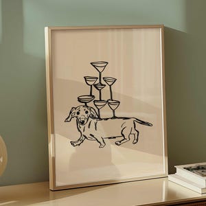 May include: A framed print featuring a black line drawing of a dachshund dog balancing a pyramid of champagne glasses on its back. The artwork has a minimalist style and is set against a neutral background.