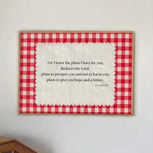 May include: A framed print with a red and white checkered border. The print features a cream-colored center with text from Jeremiah 29:11. The text is in a black, serif font. The frame is a light brown color.