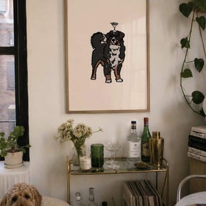 May include: A framed wall art print featuring a black, white, and brown Bernese Mountain Dog illustration with a small martini glass balanced on its head, set against a light cream background within a thin, light-colored wooden frame.