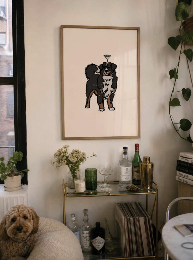 May include: A framed wall art print featuring a black, white, and brown Bernese Mountain Dog illustration with a small martini glass balanced on its head, set against a light cream background within a thin, light-colored wooden frame.