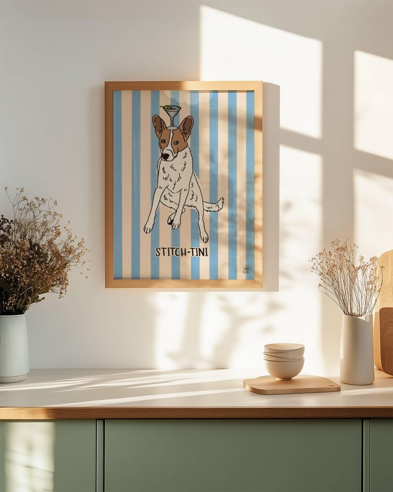 May include: A framed wall art print featuring a white and brown dog with a martini glass balanced on its head, set against a light blue and white vertical striped background with the text STITCH-TINI printed in black below the dog.
