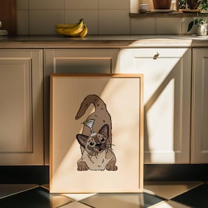 May include: A framed print featuring a tan and brown Siamese-style cat in a playful pose with a martini glass balanced on its head. The cat has dark brown ears, a dark face mask, and a long tail, set against a solid light beige background.