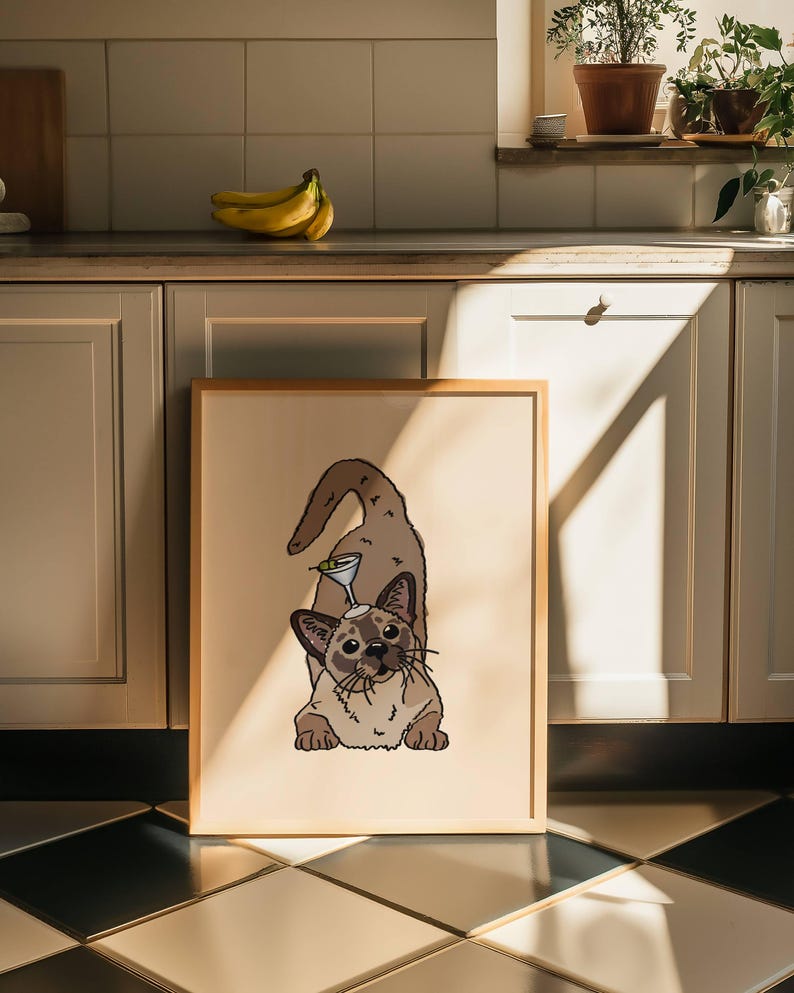 May include: A framed print featuring a tan and brown Siamese-style cat in a playful pose with a martini glass balanced on its head. The cat has dark brown ears, a dark face mask, and a long tail, set against a solid light beige background.