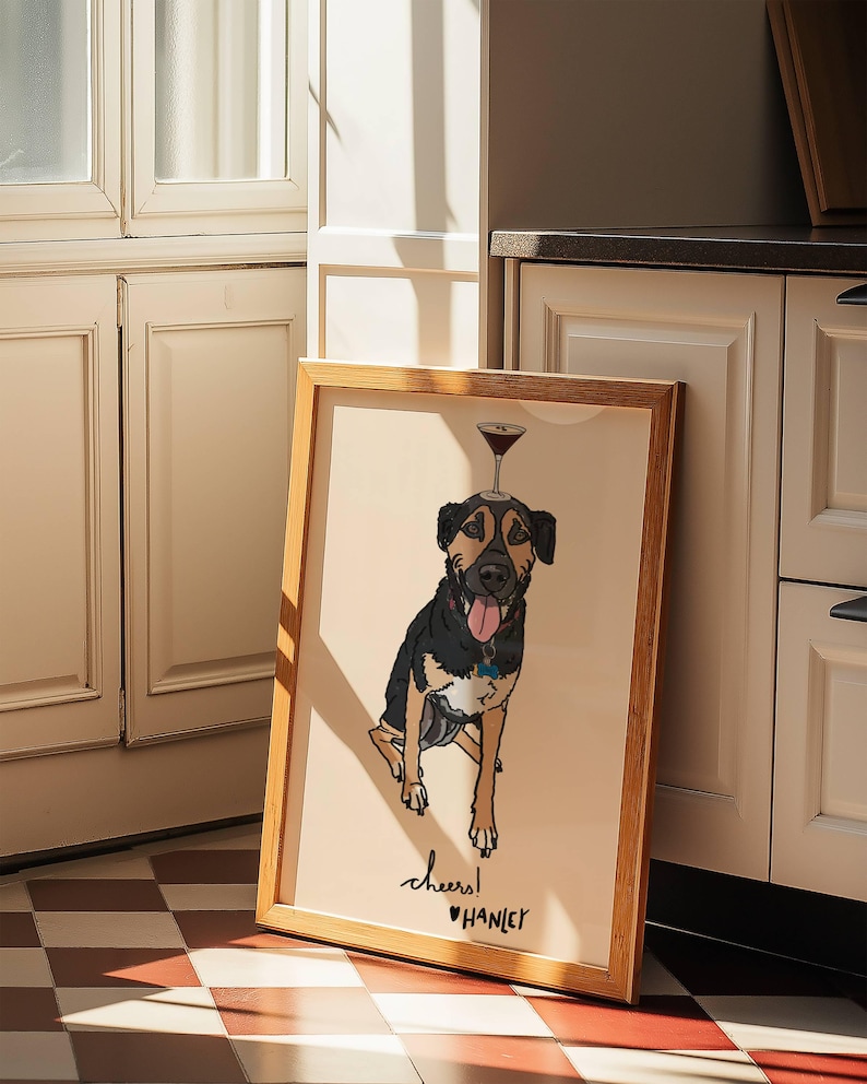 May include: A framed illustration of a black and tan dog with its tongue out, balancing a martini glass on its head. The artwork features a light beige background with the handwritten text "cheers!" and the name "HANLEY" printed below the dog.