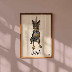 May include: A framed art print featuring a cartoon illustration of a blue heeler dog with a beer bottle balanced on its head. The dog has a grey and tan coat, a collar with a green tag, and the name LUNA is printed in black below it.