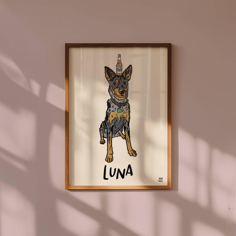 May include: A framed art print featuring a cartoon illustration of a blue heeler dog with a beer bottle balanced on its head. The dog has a grey and tan coat, a collar with a green tag, and the name LUNA is printed in black below it.