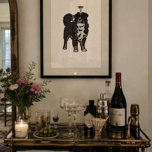 May include: A framed art print featuring a black, white, and brown Bernese Mountain Dog with a martini glass balanced on its head. The illustration is centered on a white background within a thin black frame, displayed above a bar cart.