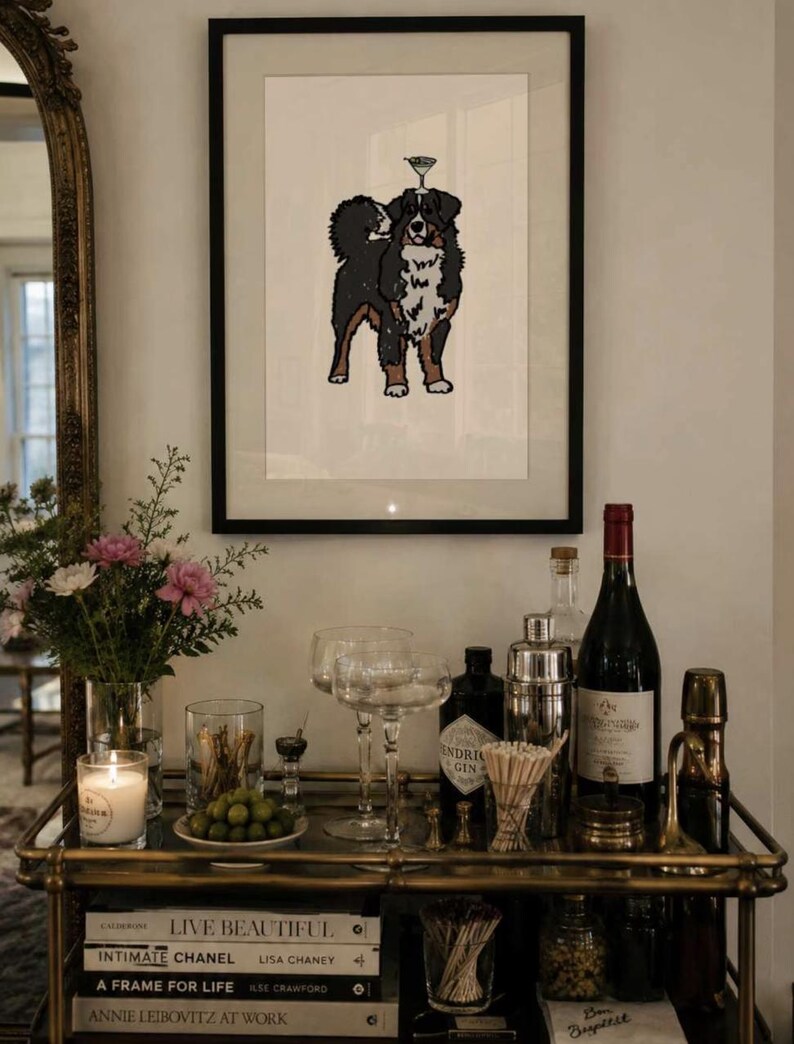 May include: A framed art print featuring a black, white, and brown Bernese Mountain Dog with a martini glass balanced on its head. The illustration is centered on a white background within a thin black frame, displayed above a bar cart.