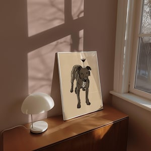 May include: A framed art print featuring a dark grey dog with a white chest patch standing on a light tan background. A small martini glass is balanced on the dog's head. The illustration uses bold black outlines and flat colours.