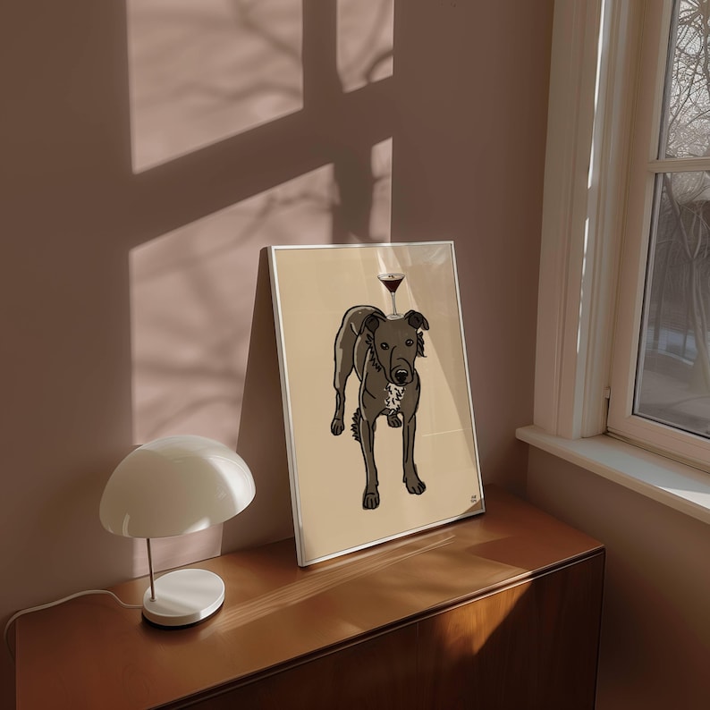 May include: A framed art print featuring a dark grey dog with a white chest patch standing on a light tan background. A small martini glass is balanced on the dog's head. The illustration uses bold black outlines and flat colours.