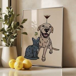 May include: A framed wall art print featuring a line-art illustration of a smiling light-coloured dog and a grey cat sitting together. The dog has a martini glass balanced on its head, and the cat has a smaller cocktail glass on its head.