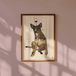 May include: A framed illustration of a brown and black dog sitting down with a wine glass balanced on its head. The artwork features a cream-coloured background and is enclosed in a simple wooden frame. The artist's signature is in the corner.