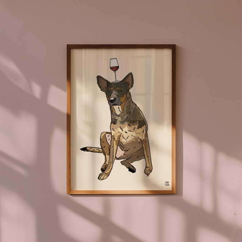 May include: A framed illustration of a brown and black dog sitting down with a wine glass balanced on its head. The artwork features a cream-coloured background and is enclosed in a simple wooden frame. The artist's signature is in the corner.