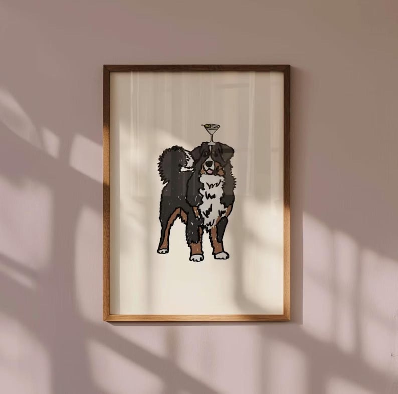 May include: A framed art print featuring a stylised illustration of a Bernese Mountain Dog with black, white, and brown fur. The dog is standing and has a small martini glass balanced on its head, rendered in simple black outlines.