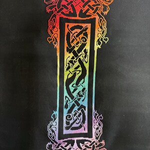 May include: A cross-stitch embroidery of the letter I on a black fabric background. The letter features a complex Celtic knotwork pattern filled with a vibrant rainbow gradient of red, orange, yellow, green, blue, and purple thread colours.