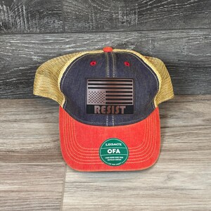 May include: A navy blue and red baseball cap with a mesh back. The front of the cap features a copper-colored American flag design with the word "RESIST" underneath. The cap has a curved brim and a small red button on top.