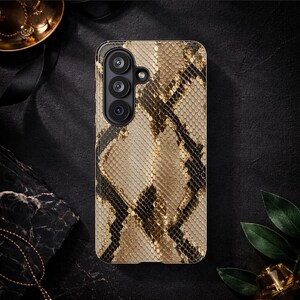 Può includere: Custodia per telefono con motivo a pelle di serpente nei toni del beige, marrone e nero. La custodia ha un modulo fotocamera con tre obiettivi. Lo sfondo è scuro, con gioielli dorati e un elemento in marmo.