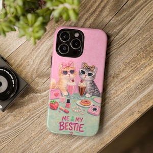 May include: Pink phone case with a cartoon illustration of two cats wearing sunglasses and bows, drinking milkshakes. The case has the text "ME & MY BESTIE" and includes small illustrations like a strawberry and a camera.