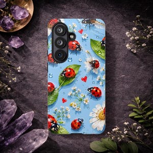 Puede incluir: Una funda de smartphone azul claro con un patrón de mariquitas rojas, pequeñas flores blancas, hojas verdes y diminutos corazones rojos. El diseño incluye gotas de agua dispersas y una abertura para una cámara de tres lentes.