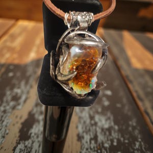 Puede incluir: Colgante artesanal con una gema iridiscente única, engastada en un marco de metal plateado. El colgante cuelga de un cordón de cuero marrón. La gema muestra una mezcla de tonos naranja, blanco y verde, creando un efecto visual llamativo.