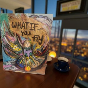 Puede incluir: Una pintura artística con el texto "WHAT IF YOU FLY". La obra presenta un águila colorida con las alas extendidas, pintada en tonos dorados, azules, verdes y morados. La pintura se exhibe sobre una mesa de madera.