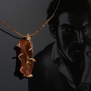 Könnte beinhalten: Eine einzigartige Anhänger Halskette mit einem grob geschnittenen, bernsteinfarbenen Stein mit silbernen Drahtakzenten. Der Stein hängt an einer zarten Goldkette. Der Hintergrund ist ein Schwarz-Weiß-Porträt.