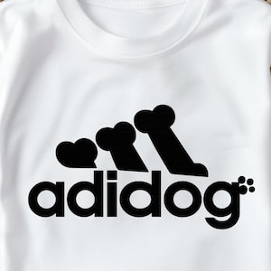 Puede incluir: Camiseta blanca con un gráfico negro que dice "adidog". El gráfico presenta tres formas de hueso en fila, asemejándose a un logotipo. La camiseta está doblada sobre una cesta tejida.