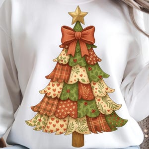May include: White sweatshirt featuring a colorful Christmas tree design. The tree is adorned with a gold star, a red bow, and patchwork-style sections in green, orange, and cream. A festive and cozy holiday-themed garment.