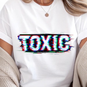 May include: White t-shirt featuring the word "TOXIC" in a glitch effect. The text is rendered in white, red, and blue, set against a black background. A casual, everyday garment.