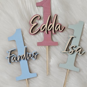 Op de afbeelding: Drie houten taarttoppers, elk met een groot cijfer één in pastelblauw, roze of saliegroen. Elke topper heeft een naam in cursief schrift: Fardus, Edda of Isa. De toppers zijn bevestigd op dunne, puntige houten stokjes voor in de taart.