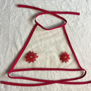 Könnte beinhalten: Ein durchsichtiges, dreieckiges Oberteil mit roter Einfassung und Bindebändern. Zwei rote Blumenapplikationen mit goldenen Zentren sind auf der Vorderseite angebracht. Der Stoff ist weiß mit kleinen, gleichmäßig verteilten weißen Punkten.