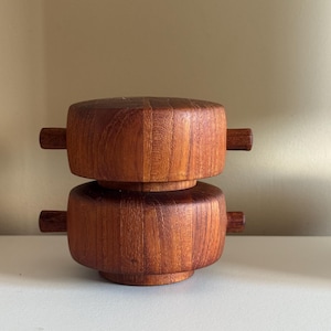 May include: Two-piece wooden container with a warm, reddish-brown finish. The top and bottom sections are cylindrical with small, rectangular handles extending from the sides. The container is likely for storage or display.