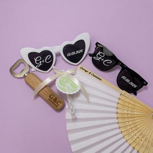 Peut inclure: Lunettes de soleil blanches en forme de cœur, lunettes de soleil noires, un décapsuleur en bois, un éventail blanc et un tube à bulles. Les lunettes portent le texte G+C et 05.09.2026. Le décapsuleur est gravé G+C 05.09.2026. L'éventail affiche George & Catalina.