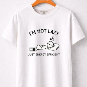 Puede incluir: Camiseta blanca con la frase negra «I'M NOT LAZY JUST ENERGY EFFICIENT». El diseño muestra un muñeco de palo durmiendo y un icono de batería baja. La camiseta está colgada en una percha naranja.