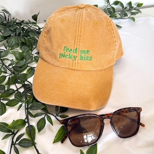 May include: A mustard yellow baseball cap with a distressed fabric finish and the embroidered green text "feed me picky bits" on the front panel, accompanied by a pair of brown tortoiseshell sunglasses with dark tinted lenses.