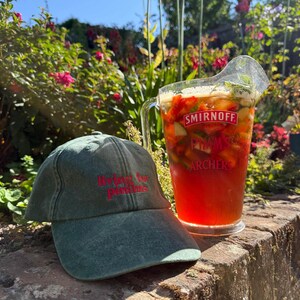 May include: A faded green baseball cap with the red embroidered text "living for pimms" sits next to a clear glass pitcher filled with a red liquid, sliced strawberries, cucumber pieces, and mint leaves, featuring the brand names Smirnoff, Pimms, and Archers.