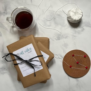 May include: An overhead shot features a clear glass mug with dark liquid, brown wrapped packages tied with black ribbon, a cork coaster with red dots, and a ball of white string. The packages have labels with text.