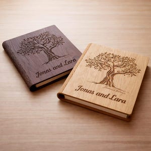 Könnte beinhalten: Zwei rechteckige Fotoalben aus Holz mit weißen Seiten. Ein Album ist aus dunkelbraunem Holz, das andere aus hellem Holz. Beide haben ein lasergraviertes Baummotiv und den Text "Jonas and Lara" auf dem Einband, die Holzmaserung ist sichtbar.
