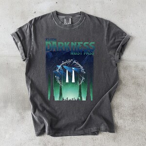 Op de afbeelding: Donkergrijs t-shirt met een grafische print. Het ontwerp toont de tekst "THE DARKNESS MUST PASS" boven een cirkelvormig element met tekst en twee witte figuren, tegen een blauwgroene achtergrond. Het t-shirt heeft een ronde hals en korte mouwen.