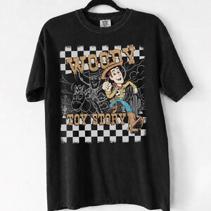 May include: A black short-sleeve t-shirt featuring a graphic print of the character Woody from Toy Story. The design includes a black and white checkered border, a desert background, and the text WOODY TOY STORY in a tan, distressed font style.
